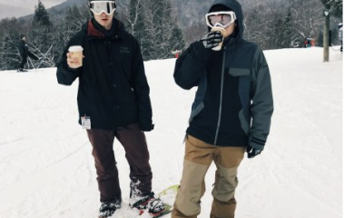 Two male snowboarders standing by snowboards on a New England mountain drinking hot chocolate