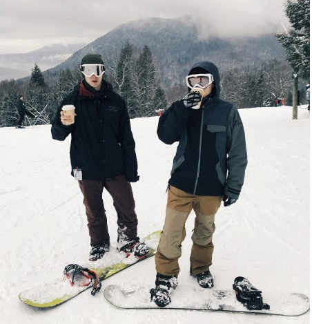 Two male snowboarders standing by snowboards on a New England mountain drinking hot chocolate