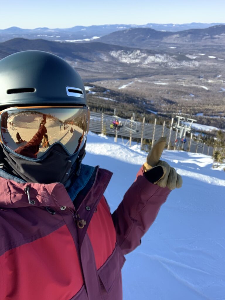 Snowboarder wearing a purple and pink jacket, black helmet, and goggles standing on a trail and pointing left hand