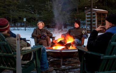 Four adults sitting around a fire pit in the winter when there is snow on the ground