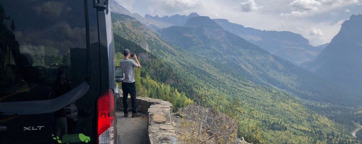 A man standing outside of a Greenvans 15 passenger van taking pictures of the mountains