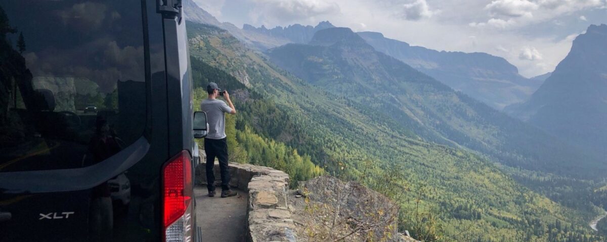 A man standing outside of a Greenvans 15 passenger van taking pictures of the mountains