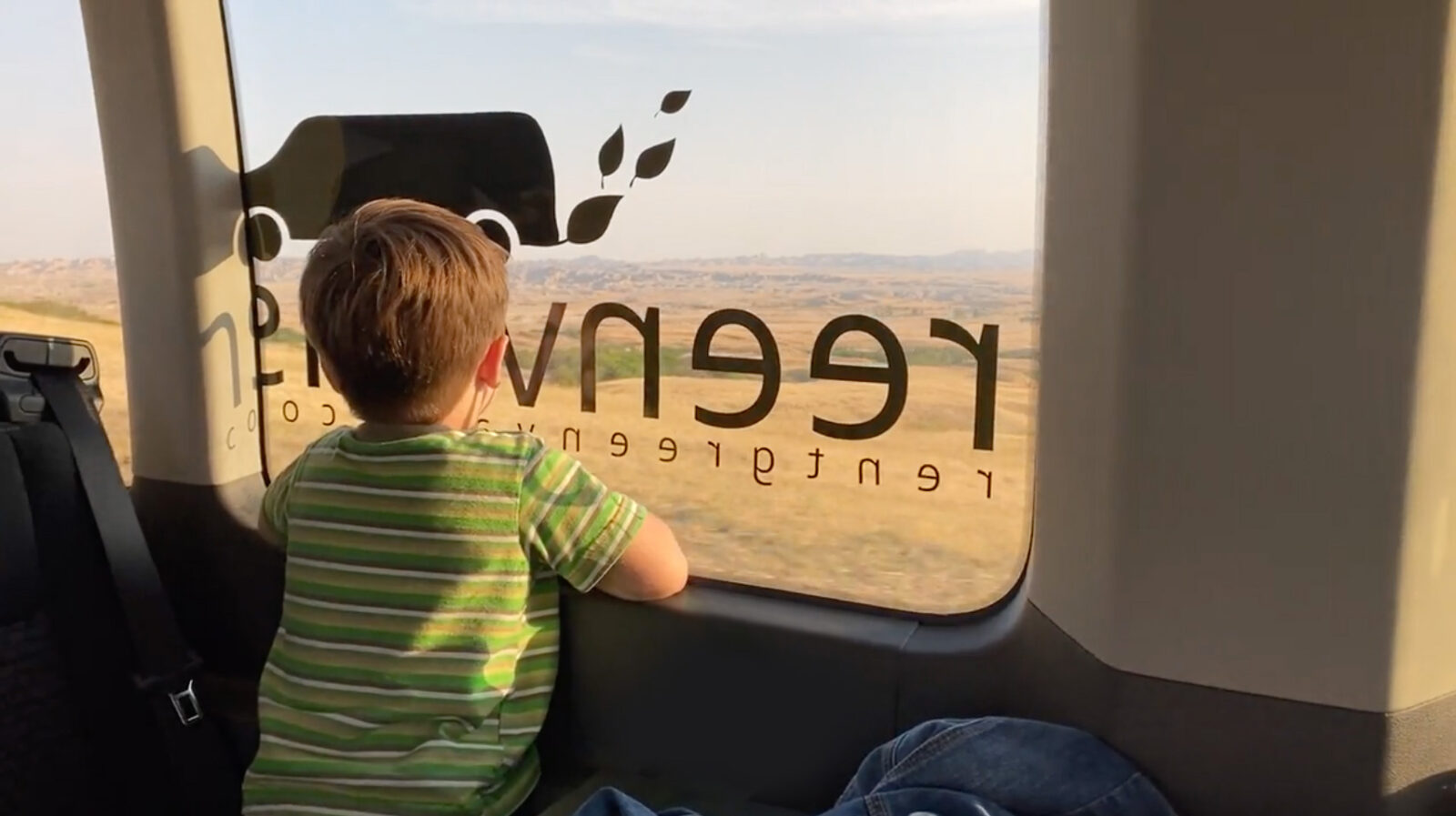 A boy looks out of a window of a Greenvans passenger van that has the Greenvans logo